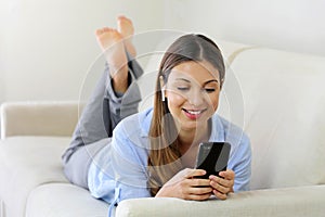 Happy relaxed young woman wearing wireless earphones listening to music lying on a couch in the living room at home