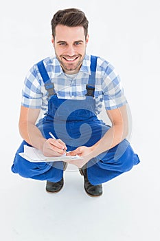 Happy repairman crouching while writing on clipboard