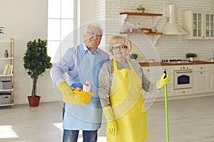 Happy retired couple in aprons and gloves standing with equipment for home cleaning