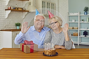 Happy retired couple in festive hats sitting with birthday cake and present box