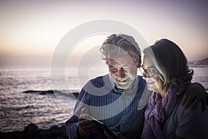 Happy retired couple having fun sitting on the cliffs, dusk light looking at the same tablet. Happy retirement concept. Sea in the