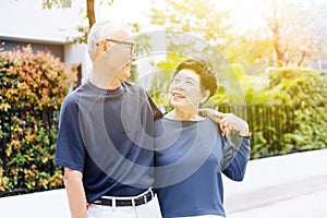 Happy retired senior Asian couple walking and looking at each other with romance in outdoor park and house in background.