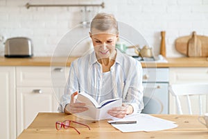 Happy retired woman sitting at kitchen and enjoying reading book