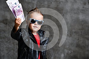 Happy rich kid boy millionaire in fashion sunglasses hold stack of 500 euro money cash in leather jacket and red t-shirt photo