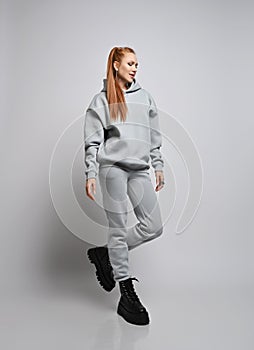 Happy rich slim red-haired woman in trendy grey sportswear hoodie, pants and black shoes walks, looking down