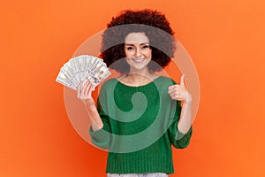 Happy rich woman with Afro hairstyle wearing green casual sweater holding big sum of money, looking