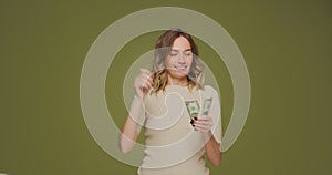 Happy rich young woman scattering dollars, throwing around cash, squandering, wasting money on green studio background