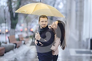 Happy romantic couple, guy and his girlfriend dressed in casual clothes are hugging under the umbrella and look at each