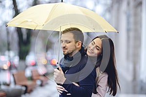 Happy romantic couple, guy and his girlfriend dressed in casual clothes are hugging under the umbrella and look at each