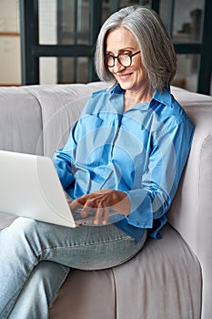 Happy 60s mature woman using laptop computer sitting on couch at home.