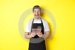 Happy salesman in black apron, holding digital tablet and looking surprised, standing against yellow background