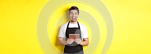 Happy salesman in black apron, holding digital tablet and looking surprised, standing against yellow background