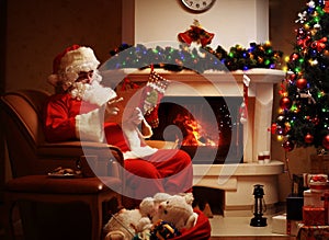 Happy Santa Claus sitting at his room at home near Christmas tree and big sack and reading Christmas letter or wish list