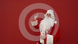 Happy Santa stands on a red background, smiling and pointing to the side on copy space. A man in a santa costume shows a finger to
