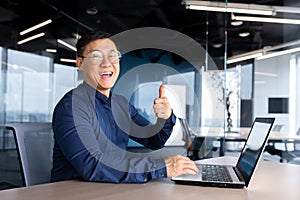 Happy and satisfied asian programmer looking at camera and smiling holding thumb up, businessman working inside office