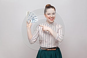 Happy satisfied attractive young businesswoman in striped shirt standing, holding many dollars, pointing finger and looking at
