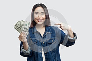 Happy satisfied beautiful young businesswoman in blue denim jacket standing, holding many dollars, pointing finger and looking at
