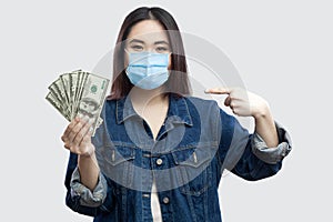Happy satisfied beautiful young businesswoman with medical mask in blue denim jacket standing, holding many dollars, pointing