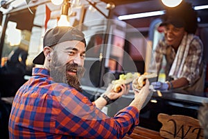 Happy, satisfied caucasian beardy male customer taking  sandwiches from a polite employee in fast food service, looking at camera