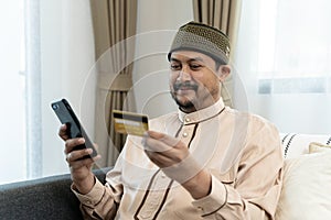 Happy and satisfied man shopping online sitting on sofa at home, hispanic customer holding bank credit card and phone