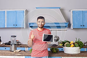 Happy satisfied man standing in kitchen, holding tablet and showing thumbs up like gesture, recommending food recipes from