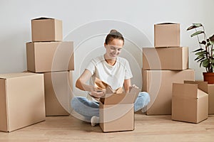 Happy satisfied woman wearing white t shirt sitting on floor surrounded with cardboard boxes with belongings, unpacking carton