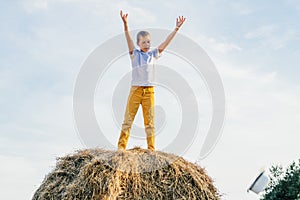 Happy school aged boy joyfully stand at top of haystack with raised arms. Carefree child on haymaking. Summer vacation.