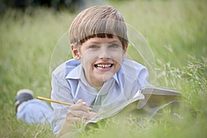Happy school boy doing homework and smiling, lying on grass
