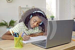 Happy school child sitting at desk with laptop, having online class and writing in notebook