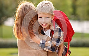 Happy schoolboy with backpack hugging embracing his mother before going to school on sunny day