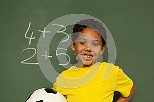 Happy schoolboy with football looking at camera against greenboard in a classroom