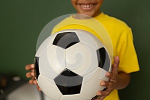 Happy schoolboy with football looking at camera against greenboard in a classroom
