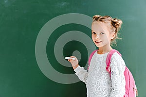 happy schoolchild holding chalk near chalkboard on green .