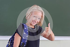 Happy schoolgirl with backpack showing thumb up in a classroom near green chalkboard. The child likes to learn. Back to school.