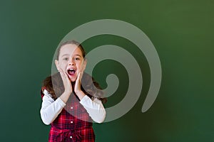 Happy schoolgirl preschool girl in plaid dress standing in class near a green blackboard. Concept of school education