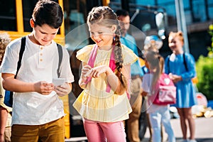 happy schoolgirl and schoolboy using smartphone together in front of school bus