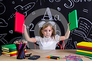 Happy schoolgirl sitting at the desk with books, school supplies, holding up two books in her both arms