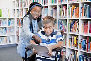 Happy schoolgirl standing with schoolboy on wheelchair using digital tablet
