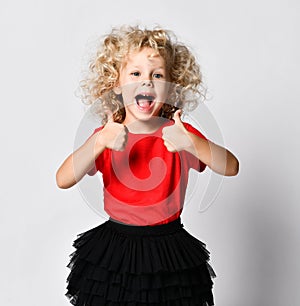 Happy screaming curly hair blonde kid girl in red t-shirt and black skirt shows thumbs-up with both hands