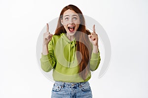 Happy screaming girl with red hair, pointing fingers up and looking amazed at promo store advertisement, checking out