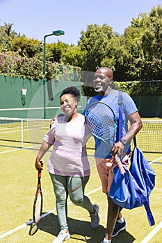 Happy senior african american couple walking with rackets on sunny grass tennis court, copy space