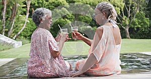 Happy senior african american female friends making a toast with drinks sitting by pool, slow motion