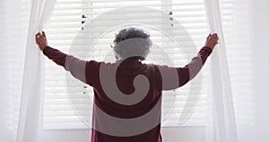 Happy senior african american woman widening curtains in bedroom