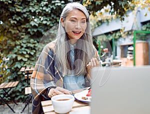 Happy senior Asian lady eats toast with strawberries watching video via laptop at table outdoors