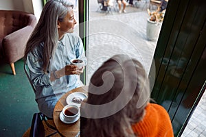 Happy senior Asian woman with friend look through window sitting at table in cafe