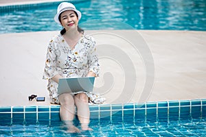 happy Senior asian woman working on  laptop computer sitting at poolside with put her legs in the water . relaxation retirement