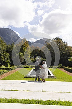 Happy senior biracial bride and groom dancing in garden at sunny wedding ceremony, copy space