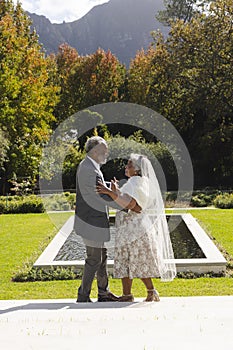 Happy senior biracial bride and groom dancing in garden at sunny wedding ceremony, copy space