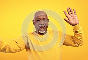 Happy senior black man taking selfie, waving at camera on orange studio background