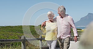 Happy senior caucasian couple with arms around each other walking on path leading to the beach